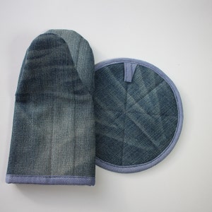 May include: A denim oven glove and circular pot stand set. The oven glove is a standard shape, and the pot stand is round with a small hanging loop. Both items are made from blue denim fabric with a blue and white trim.