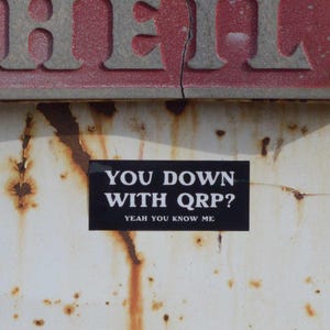 Könnte beinhalten: Ein verwittertes Metallschild mit dem Wort "HELL" in silbernen Buchstaben. Darunter steht auf einem rostigen, weißen Untergrund ein schwarzer Aufkleber mit der Aufschrift "YOU DOWN WITH QRP? YEAH YOU KNOW ME".