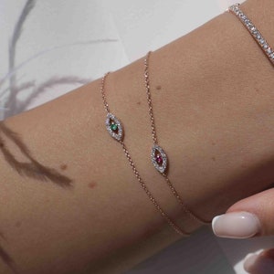 Könnte beinhalten: Das Handgelenk einer Frau ist mit drei zarten Armbändern geschmückt. Zwei zeigen ein kleines Augenmotiv mit einem grünen und roten Edelstein, während das dritte ein schlichtes, mit Diamanten besetztes Band ist. Ihre Finger sind mit zwei Ringen geschmückt, einer mit einem mit Diamanten besetzten Band und der andere mit einem mit Diamanten besetzten Design.