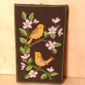 Op de afbeelding: Een bruine wandhanger van stof met een kruissteekontwerp van twee gele vogels die op een tak zitten met roze bloemen.
