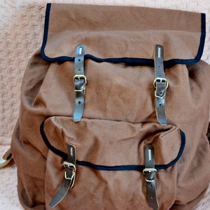 May include: A brown canvas backpack with black trim and leather straps and buckles. The backpack has two large pockets on the front.