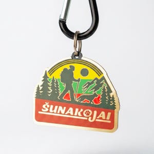 May include: A wooden keychain with a black carabiner clip. The keychain features a colorful design of a hiker, dog, mountains, and trees, with the text "ŠUNAKOJAI" in white letters on a red background.