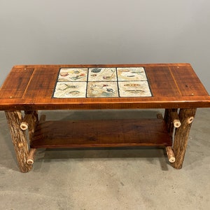 May include: A rustic wooden coffee table with a lower shelf. The top of the table features a decorative tile inset with a design of various fishing lures.