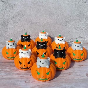 May include: A collection of orange Halloween pumpkin figurines with carved faces. Each pumpkin has a small cat figurine on top, with some cats black and some white. The pumpkins have green stems and sit on a light-colored surface.