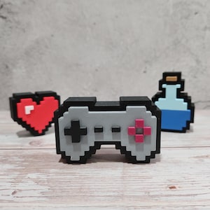 May include: Pixelated game-themed figurines. A gray and black controller with pink and black buttons is centered. A red heart with a white pixel is to the left. A blue potion bottle with a brown top is to the right. All are on a light wood surface.