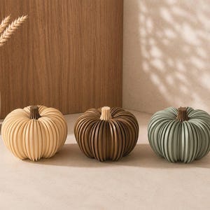 May include: Three decorative pumpkins with a ribbed, layered design. The left pumpkin is light beige with a dark brown stem, the center pumpkin is dark brown with a light beige stem, and the right pumpkin is sage green with a dark brown stem.