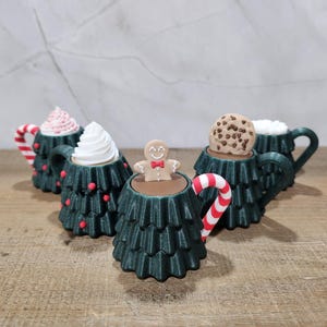 May include: A collection of Christmas tree-shaped mugs, each filled with a different topping. The mugs are dark green with red accents and candy cane handles. Toppings include whipped cream, a gingerbread man, and a chocolate chip cookie.