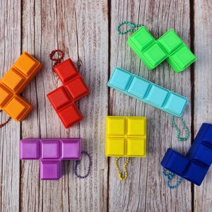 May include: A collection of colorful, geometric puzzle pieces in orange, red, green, blue, yellow, and purple. Each piece has a keychain attached. The pieces are arranged on a light brown wooden surface.