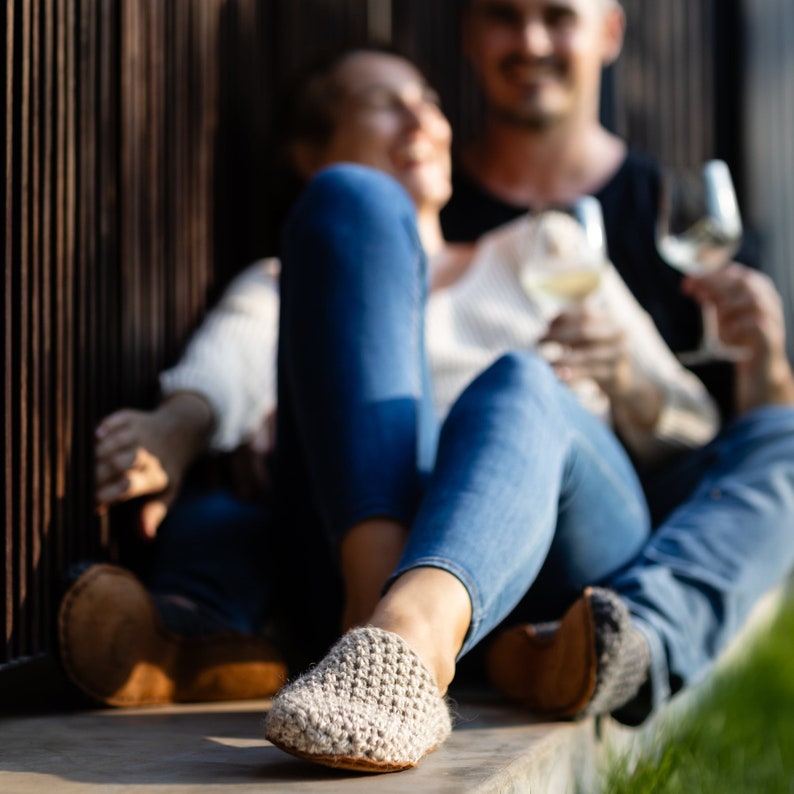 A pair of light gray crochet slippers with a leather sole. The slippers are worn by a person wearing blue jeans. A second person is in the background, holding a glass of white wine.