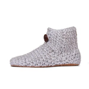 A light gray hand knitted slipper boot with a wooden button closure. The slipper has a thick, textured knit and a leather sole. Barefoot slippers for indoor use. Warm, cozy and comfortable.