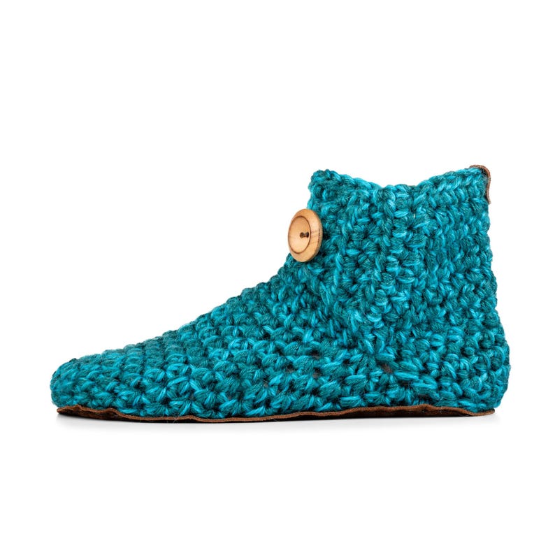A turquoise blue hand knitted slipper boot with a wooden button closure. The slipper has a thick, textured teal knit and a leather sole. Barefoot slippers for indoor use. Warm, cozy and comfortable.