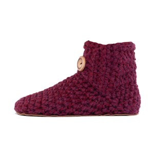 A burgundy red crochet slipper boot with a wooden button closure. The slipper has a thick, textured wool appearance and a leather sole. The boot has a rounded toe. The warm and cozy footwear is designed for indoor use and is a comfortable option for lounging at home.