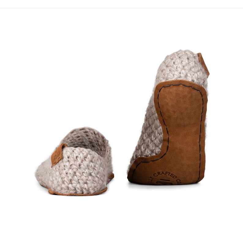 Peut inclure: Deux chaussons de b&eacute;b&eacute; en crochet gris clair avec des semelles et des accents en cuir marron. Les chaussons sont faits &agrave; la main et pr&eacute;sentent un motif tricot&eacute; textur&eacute;. Un chausson est droit, l'autre est couch&eacute; sur le c&ocirc;t&eacute;.