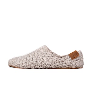 A light beige knitted slipper with a brown leather label on the heel. The slipper has a rounded toe and a suede sole. The knitted texture is visible throughout the slipper.