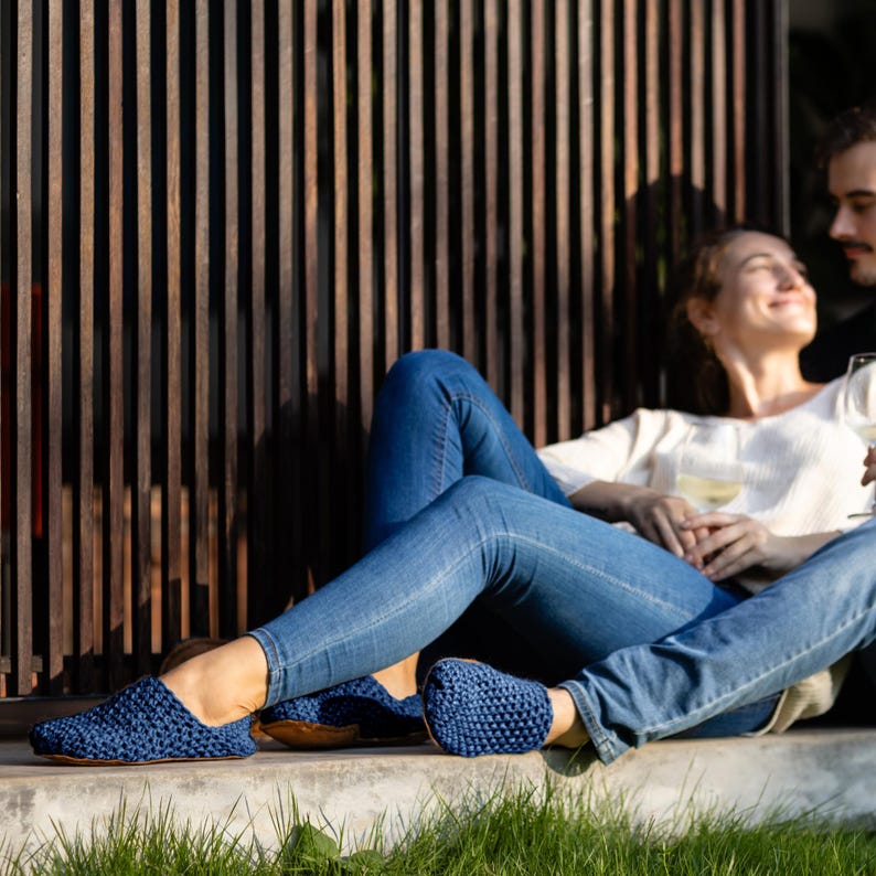 A pair of navy blue knitted slippers. The slippers are made of a textured knit material and have a rounded toe. The slippers are worn by a person wearing blue jeans.