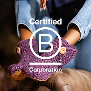 A pair of purple crocheted slippers with wooden buttons and tan leather soles. The image includes the text "Certified B Corporation" in white, within a white circle. Kingdom of Wow is proudly B Corp Certified
