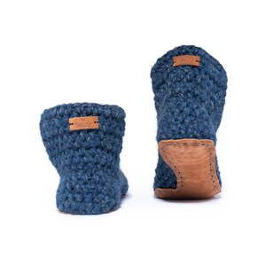 A navy blue hand knitted slipper boot with a wooden button closure. The slipper has a thick, textured knit and a leather sole. Barefoot slippers for indoor use. Warm, cozy and comfortable. Back view showing handstitched suede sole and labelling
