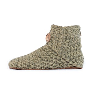 A olive green knitted slipper boot with a wooden button closure. The slipper has a thick, textured wool knit and a leather sole. The boot has a rounded toe. The warm and cozy footwear is designed for indoor use and is a comfortable option for lounging at home.