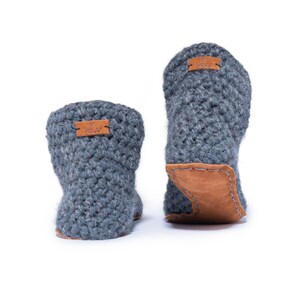 A dark gray crocheted slipper boot with a wooden button closure. The boot has a rounded toe and a leather sole. The warm and cozy footwear is designed for indoor use and is a comfortable option for lounging at home. Back view showing off the handstitched leather sole
