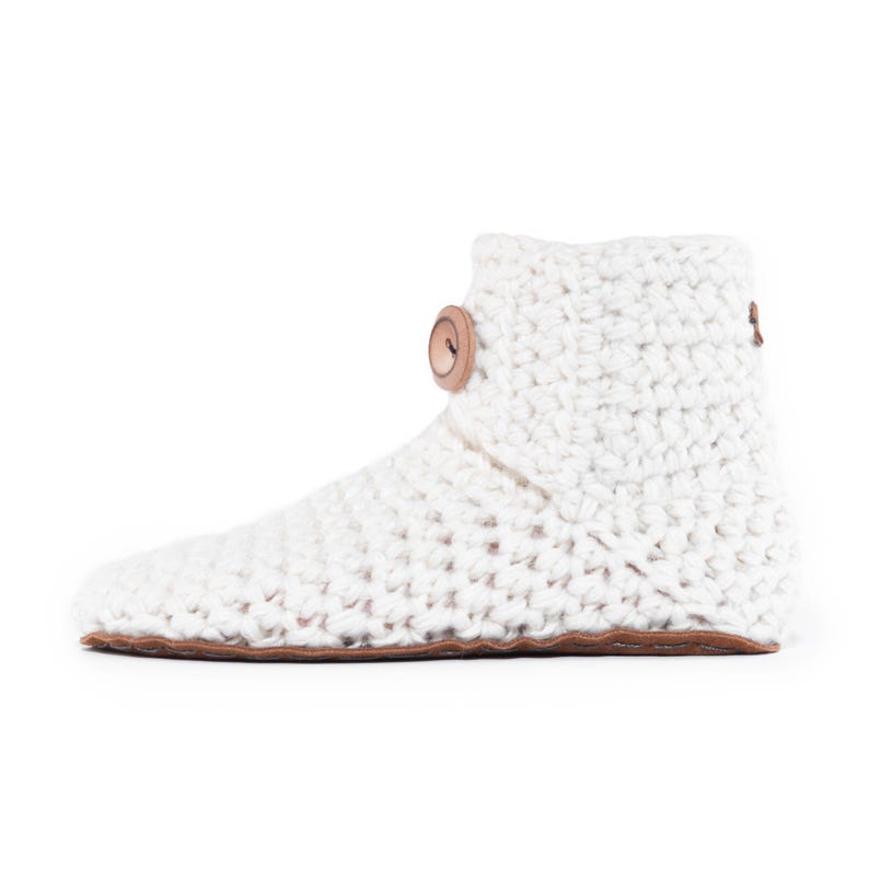 A white, crocheted slipper boot with a suede leather sole and a large, round, wooden button closure. The boot is made of thick, textured wool yarn and has a high ankle. The overall style is cozy and handmade. Stylish and hygge.