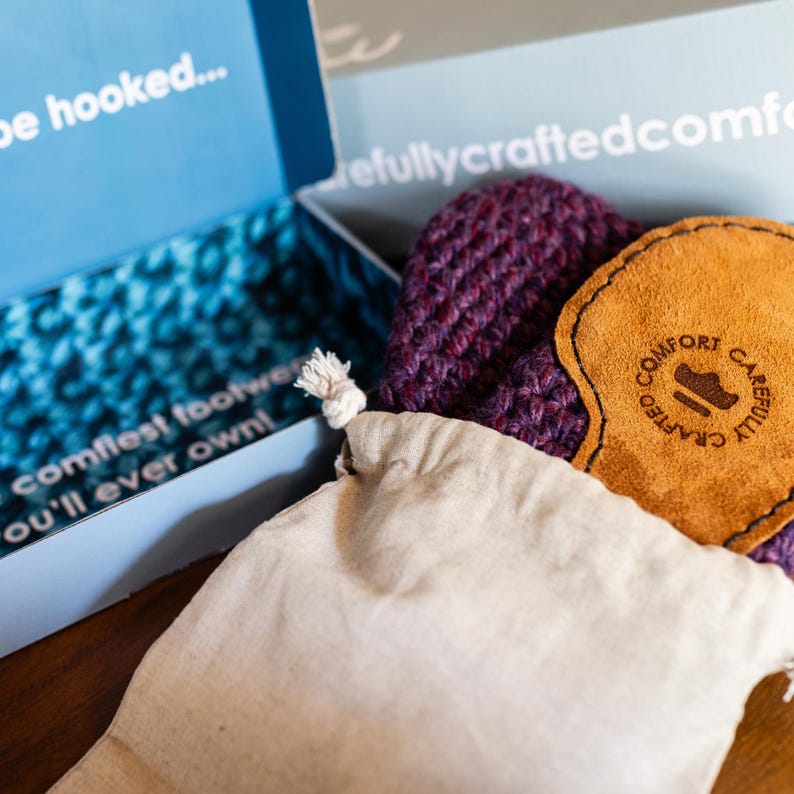 A pair of purple knitted slippers with a leather insole, presented in a blue packaging box and cotton drawstring bag. The box has text that reads "comfort footwear you'll ever own!"
