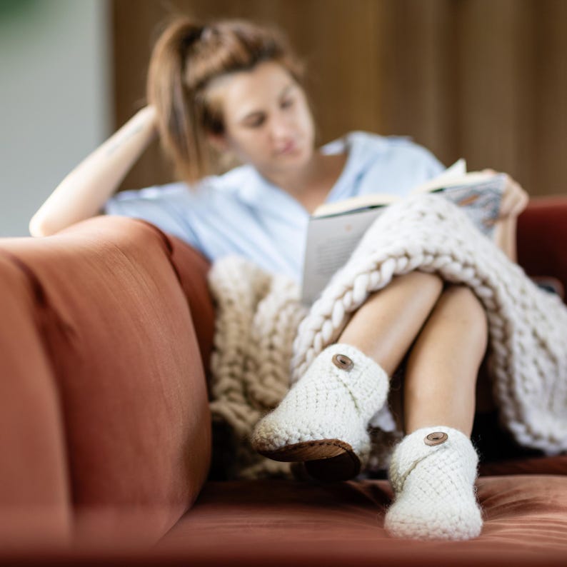 Woman sits on a rust red sofa reading a magazine in a cozy setting. She wears a blanket over her knees and white boot wool slippers with a button closure and soft leather soles.