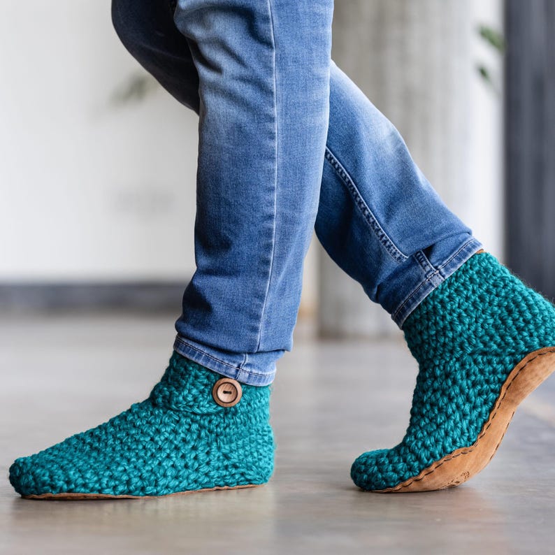 A man's feet wears teal colored knitted wool slippers with button closure and leather soles with blue denim jeans