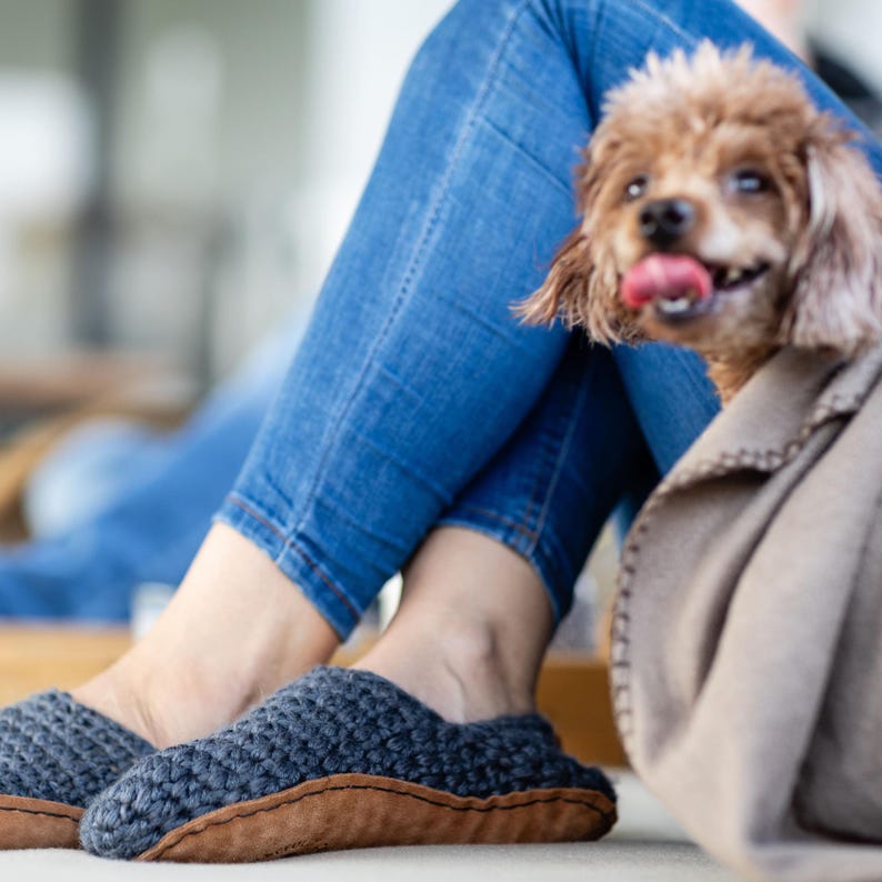 A pair of dark gray knitted slippers with suede leather soles. The slippers are worn by a person wearing blue jeans. A small brown dog with its tongue out is in the background.