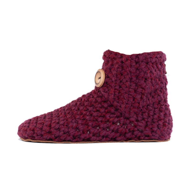 A mulberry red hand knitted slipper boot with a wooden button closure. The slipper has a thick, textured knit and a leather sole. Barefoot slippers for indoor use. Warm, cozy and comfortable. Perfect loungewear.