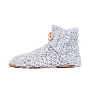 A light gray crocheted slipper boot with a wooden button closure. The boot has a thick, textured knit and a brown sole. The cozy ankle-high boot is designed for indoor wear and comfort.