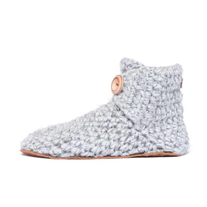 A light gray crocheted slipper boot with a wooden button closure. The boot has a thick, textured knit and a brown sole. The cozy ankle-high boot is designed for indoor wear and comfort.
