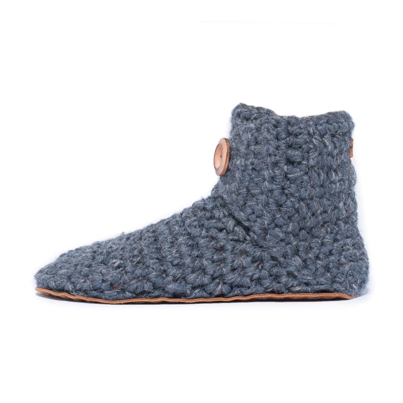 A dark gray crocheted slipper boot with a wooden button closure. The boot has a rounded toe and a leather sole. The warm and cozy footwear is designed for indoor use and is a comfortable option for lounging at home.