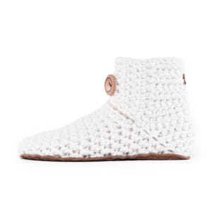 A white, crocheted slipper boot with a suede leather sole and a wooden button closure. The boot is made of thick, textured wool yarn and has a cozy, handmade appearance. The design is simple and functional, perfect for comfortable indoor wear.