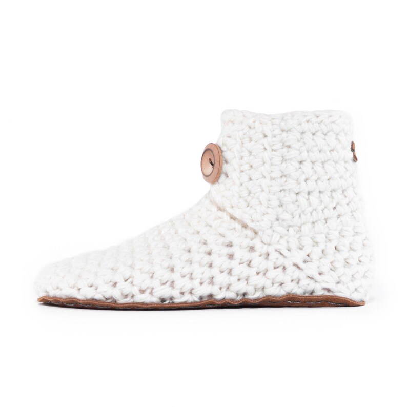 A white, crocheted slipper boot with a suede leather sole and a wooden button closure. The boot is made of thick, textured wool yarn and has a cozy, handmade appearance. The design is simple and functional, perfect for comfortable indoor wear.