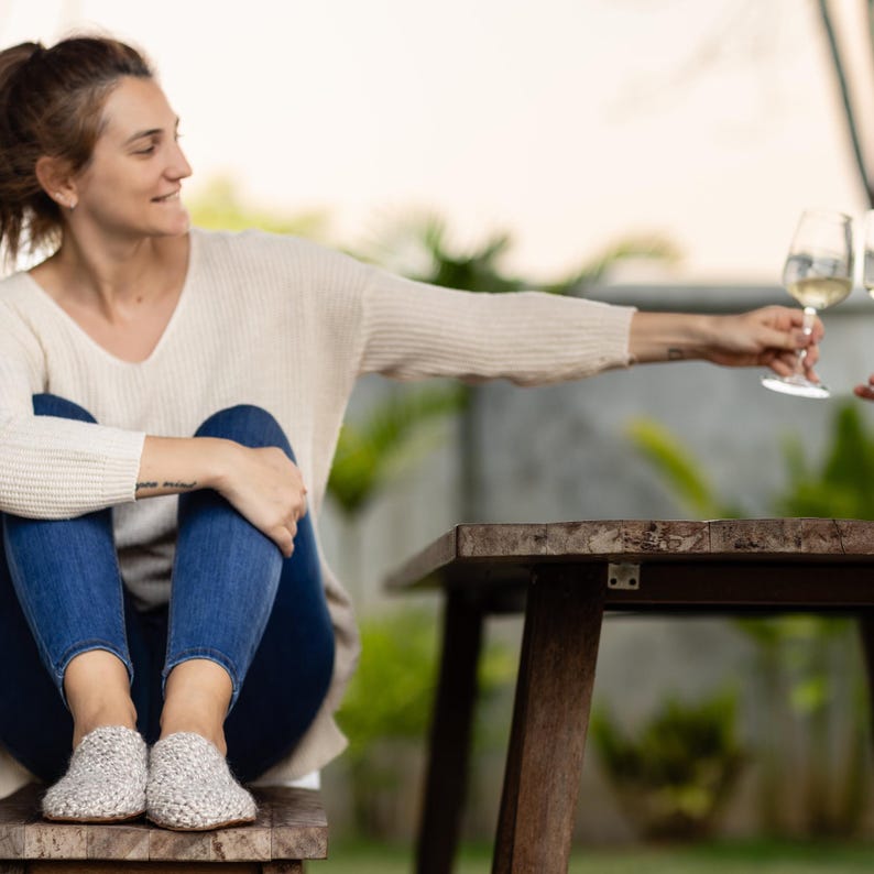 Woman sitting on a bench wearing jeans, beige knitted sweater and light beige slippers. She is clinking wine glasses with someone out of shot.