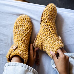 A pair of handmade, yellow crochet slippers with wooden button closures. The slippers are made of a thick, textured wool yarn and appear warm and cozy. The slippers are shown on a light blue surface.