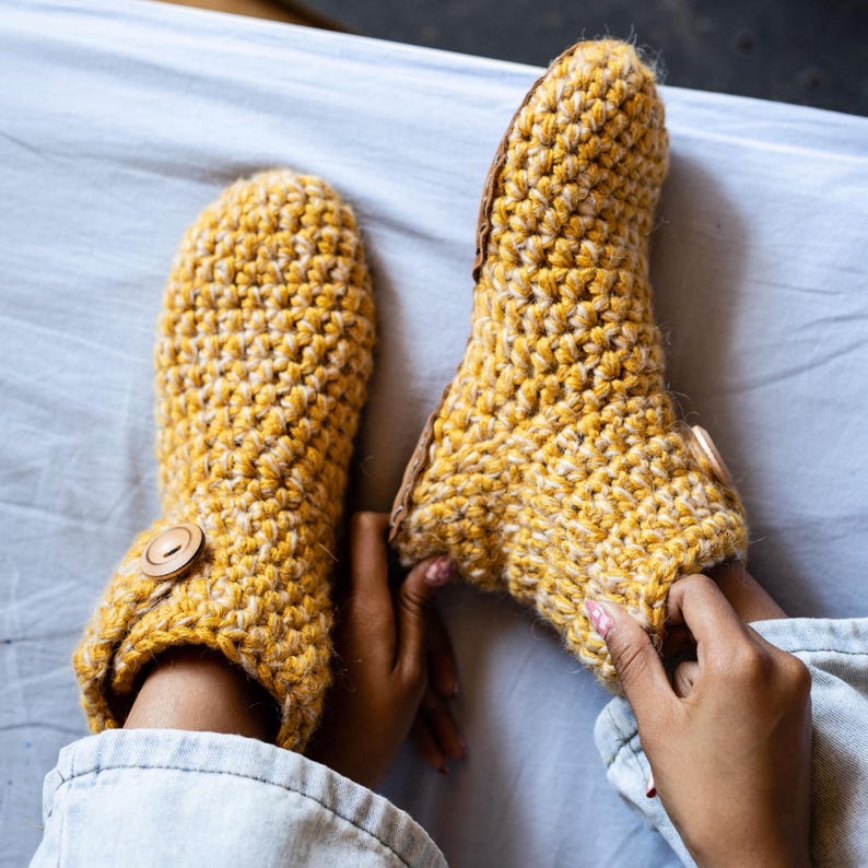 A pair of handmade, yellow crochet slippers with wooden button closures. The slippers are made of a thick, textured wool yarn and appear warm and cozy. The slippers are shown on a light blue surface.