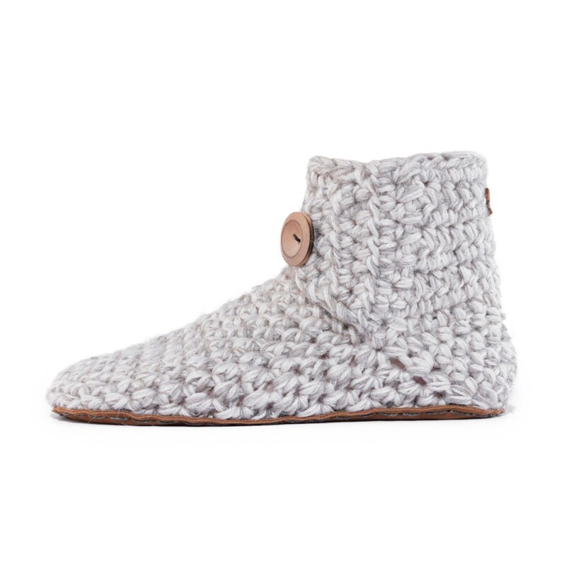 A beige off-white knitted slipper boot with a wooden button closure. The boot has a rounded toe and a leather sole. The warm and cozy footwear is designed for indoor use and is a comfortable option for lounging at home.