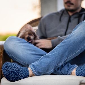 Close-up of blue knit slippers. The slippers are a deep navy blue color and handmade. The image shows the slippers on a person's feet, with blue jeans and a dog in the background.