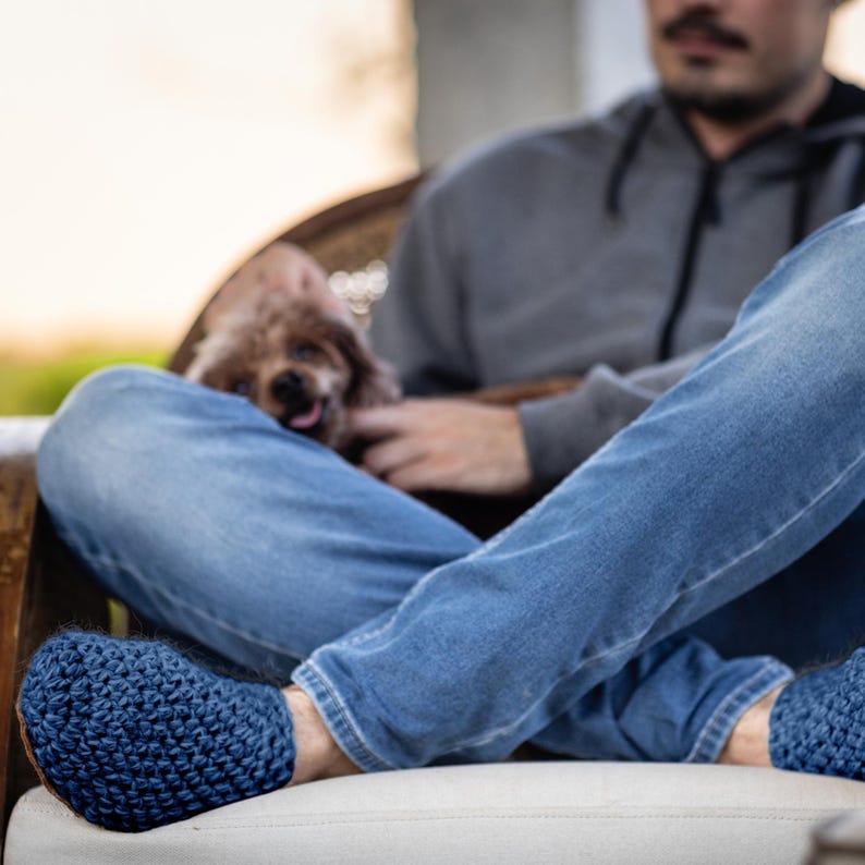Close-up of blue knit slippers. The slippers are a deep navy blue color and handmade. The image shows the slippers on a person's feet, with blue jeans and a dog in the background.