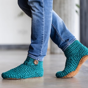 Teal crochet slippers with a button closure and a brown leather sole. The slippers are worn with blue jeans. The crochet stitch is visible, and the slippers appear warm and comfortable.