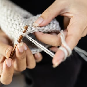 Close-up of hands crocheting with a wooden crochet hook and off-white yarn making a knitted sock. The hands are holding the hook and yarn, creating a stitch in light beige yarn.