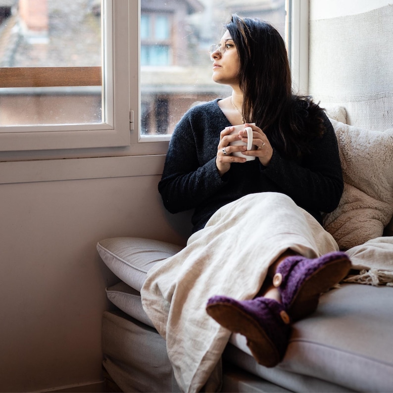 Women sits on a light colored couch holding a hot drink and looking out of a window. She is wearing a blanket and pair of purple ankle wool boot slippers. Designed for indoor use.