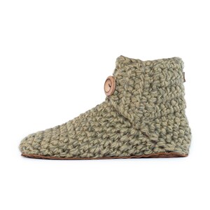 A light green, chunky-knit slipper boot with a round, wooden button closure. The boot has a leather sole and a high ankle. The cozy, handmade slipper is designed for indoor wear. Warm, cozy and comfortable. Perfect loungewear.
