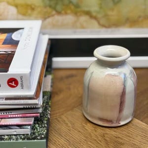 May include: A small, off-white ceramic vase with pink and blue watercolor-style accents. The vase has a rounded body and a narrow neck. A stack of books is visible to the left.
