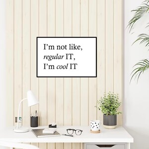 Könnte beinhalten: Ein weißer Schreibtisch mit einer weißen Lampe, einer Topfpflanze, einer Tasse mit schwarzen und weißen Punkten, einem Notizbuch, einer Brille und einem schwarzen Stift. Ein gerahmter Druck mit dem Text "I'm not like, regular IT, I'm cool IT" hängt an der Wand hinter dem Schreibtisch.