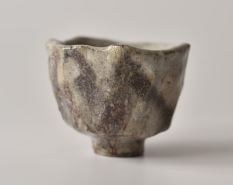 Puede incluir: Una pequeña taza de cerámica formada a mano con un borde irregular y ondulado y una base gruesa y texturizada. El exterior presenta un esmalte moteado en tonos marrón tierra, gris apagado y crema, creando una apariencia orgánica y desgastada.