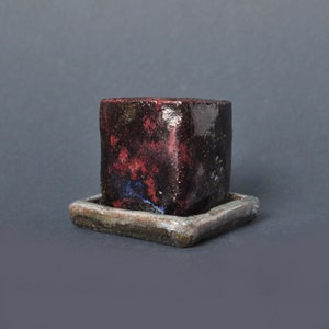May include: A small, square ceramic planter with a glossy, dark glaze featuring mottled patches of deep red, black, and hints of blue. The planter sits on a matching square ceramic saucer with a textured, muted grey and brown finish.