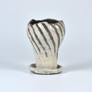 Op de afbeelding: Een kleine witte keramische pot met bruine strepen. De pot heeft een uitlopende bovenkant en een ronde basis.