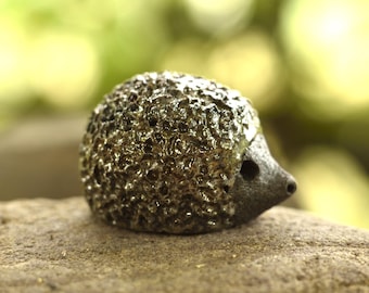 Puede incluir: Una pequeña figura de erizo esculpida que presenta un dorso oscuro, texturizado y de aspecto metálico con una superficie porosa e irregular. La cabeza es lisa, mate y de color gris oscuro, con una pequeña hendidura circular para el ojo y un hocico puntiagudo.